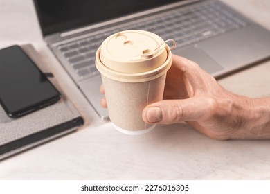 Hand holding beige paper cup, takeaway takeout coffee mug with lid at office desk, workplace closeup. - Powered by Shutterstock