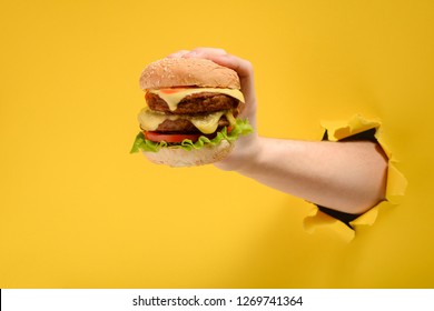 Hand holding a big burger through a hole in torn yellow paper wall. Special offer and cheap price on fast food.