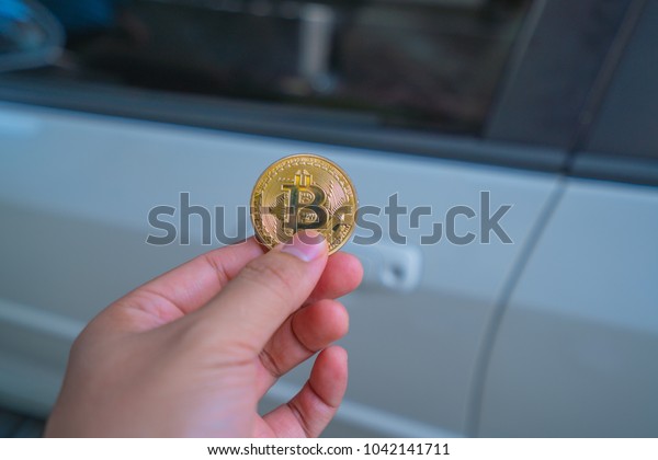 Hand holding a bitcoin in front of car. Concept\
of buying car using\
bitcoin.