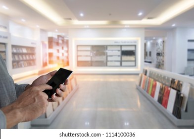 Hand Holding Black Phone With Modern Ceramic Tiles Display In The Luxury Shopping Mall. Bussiness Man Pointing Finger On Screen Smartphone On Home Material Shop Background