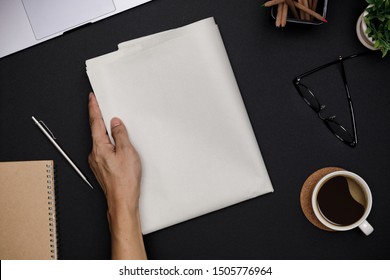 Hand Holding Blank Business Newspaper And Office Supplies With Notebook, Hot Coffee Cup, Glasses, Books And Accessories On Black Desk Topview, Daily Newspaper Mockup Concept