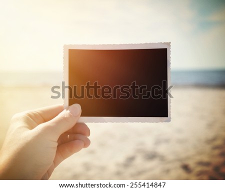 A hand is holding up a blank photo snapshot at a sunny beach to add your own photo or message in the area.