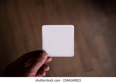 Hand Holding A Blank White Square Paper Card With Rounded Corners. Business Card, Magnet, Bumper Sticker Mock-up. Wooden Floorboard Background.