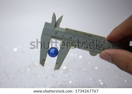 Hand holding a blue diamond with vernier caliper, tweezers and a pile of diamond. Worker measures the diameter of gems on a jewelry scale isolated on white background.