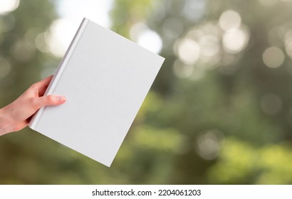 Hand Holding Book Mockup On Blurred Background With Green Trees, Grass. Literature About Nature, Ecology. Reading Leisure, Harmony Concept. High Quality Photo