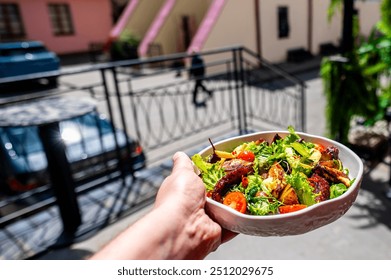 hand holding a bowl of fresh salad with mixed greens, cherry tomatoes, and grilled chicken, set against blurred outdoor balcony background. Perfect for healthy eating, lifestyle, and food-related  - Powered by Shutterstock