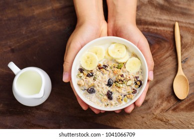 Hand Holding Bowl Of Muesli With Cereal, Slice Banana And Milk On Wooden Background, Healthy Breakfast, Top View