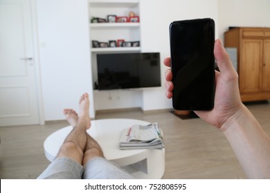 A Hand Holding A Brand New Mobile Phone With An Empty, Black Screen. The Person's Feet And Legs Are On The Living Room Table. On The Background, A Television With Black Screen Is Hanging From The Wall