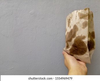 Hand Holding A Brown Paper Bag With Very Greasy Contents.  Too Much Oil Can Cause Illness In Humans. Grey Background. 