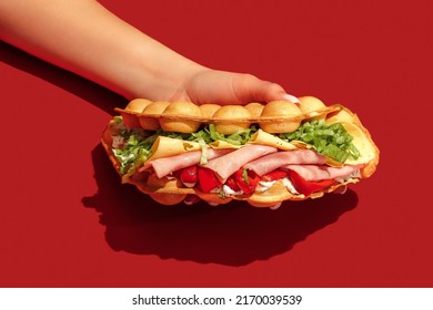Hand holding bubble waffle sandwich with ham, tomato, lettuce, bacon, sauce, mustard, cheese, red bell pepper isolated on red background. Fast food, take away. Snack