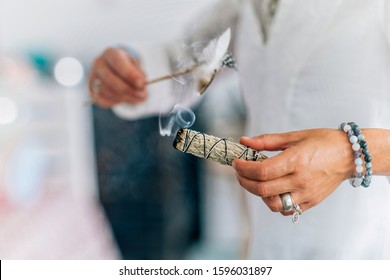 Woman’s hand holding bundle of dried sage herb, performing smudging ritual, cleansing negative energy and purifying living space with smoke