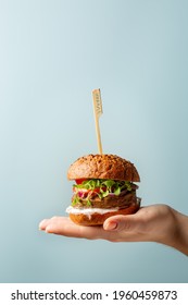 Hand Holding Burger. Hamburger With Meat Free Plant Based Cutlet, Tomatoes, Onions, White Sauce And Microgreens On Blue Background. Healthy Vegan Or Vegetarian Food Concept. Copy Space.