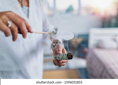 Woman’s hand holding burning sage smudge stick, spreading the smoke around with white feather, blessing and cleansing home from negative energies 