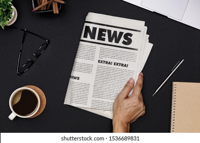 Hand Holding Business Newspaper And Office Supplies With Notebook, Hot Coffee Cup, Glasses, Books And Accessories On Black Desk Topview, Daily Newspaper Mockup Concept