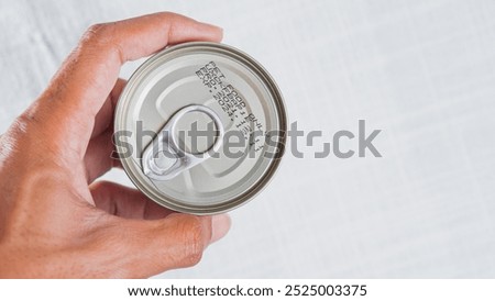 A hand holding a can with a pull tab, showing the top view of the can. The can has a metallic finish and some printed text, likely indicating the product details and expo
