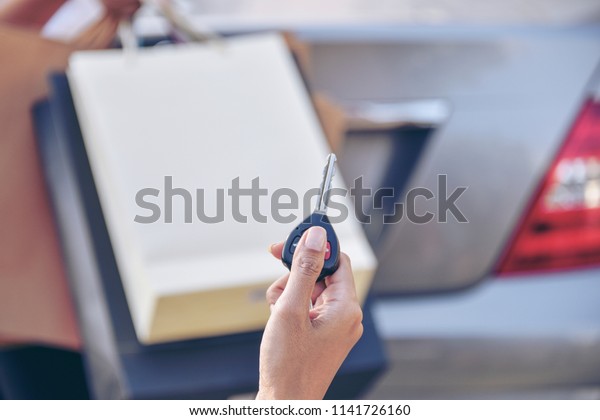 Hand holding car keys remote with blurred\
happy  young woman carrying many colourful shopping bags in the car\
park. Let’s go to shoes store\
more.