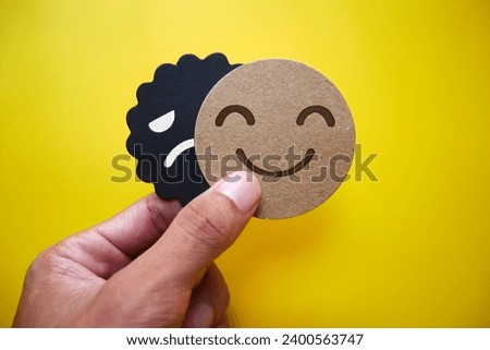 Hand holding card sad face hiding behind happy face with copy space. Concept of introversion, bipolar, depression, mental health, split personality, mood swings.