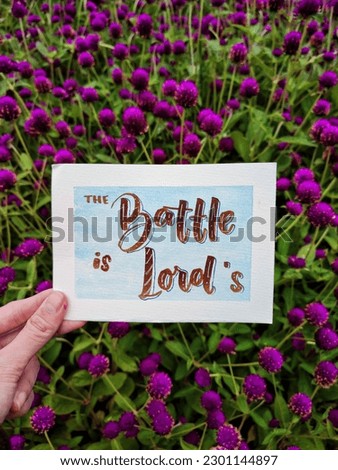 A hand holding a card written with hand-lettering style, saying The Battle is the Lord's with the pretty background of garden or flowers