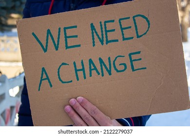 Hand Holding Cardboard Box With We Need A Change Text. Man Protesting To Environmental Pollution, Inequality, Racism Or Something Else.