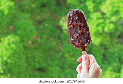 Hand Holding A Chocolate Dipped Ice Cream Bar Against  Blurry Green Foliage