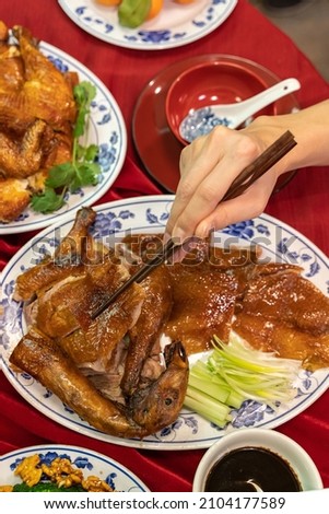 A hand holding chopsticks tries to pick up pieces of roast duck on a table filled with different Chinese food dishes for Lunar New Year. Duck symbolizes fidelity in Chinese culture.