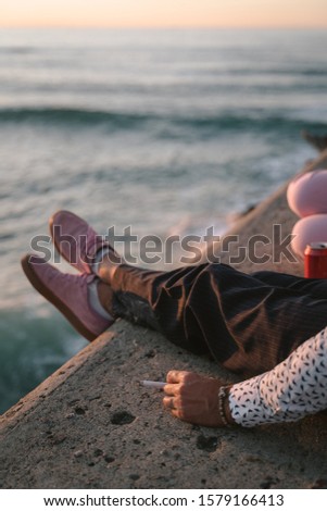 Hand holding a cigarette sitting on a stone ledge with pink shoes by the sea