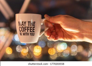 A hand holding a coffee cup is in focus with bokeh lights in the background, and has a funny quote that is a play on a famous sci-fi movie line. 