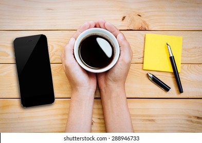 hand holding coffee cup with smartphone and notepad on wood table top view