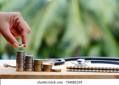 Hand Holding Coin, Stack Of Money, Stethoscope And Notebook On Wood Table, Healthcare And Business Concept