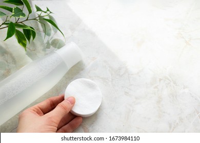 Hand Holding Cotton Pads And Make-up Remover. Toner Or Tonic For Skincare And Plant Leaves On Marble Background With Copy Space, Top View. Natural Cosmetic Concept