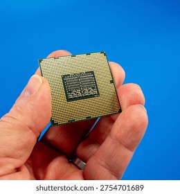 Hand holding a CPU processor showing the gold contact pins (LGA PGA socket side) against a bright blue background. Close-up macro shot of a computer central processing unit, ideal for technology, hardware, and computing concepts.