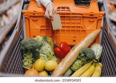 Hand holding a credit card over a shopping cart full of fresh produce in a supermarket, ready for contactless payment - Powered by Shutterstock