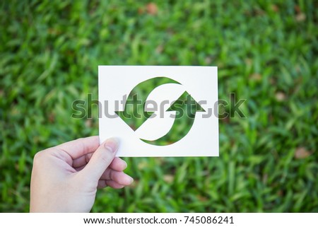 Hand holding cut paper with the logo of recycling over green grass. Recycling sign and symbol background banner concept, Earthday 