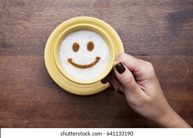 Hand holding a cute yellow cup with coffee cream. Food art creative concept image, happy face drawing with cinnamon powder over wooden background. - Powered by Shutterstock