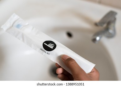 Hand Holding A Dental Kit Set Against Toilet Wash Basin. Concept Of Oral Care, Hygeine And Travel