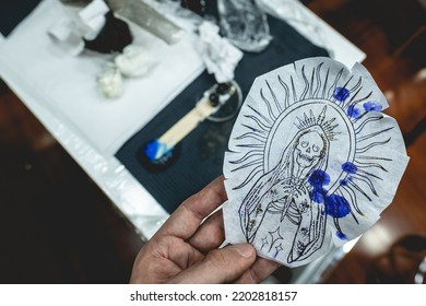 Hand Holding The Design Of A New Tattoo Of 'La Santa Muerte' (Our Lady Of Holy Death) In Tracing Paper Over The Tattoo Artist Table With Tools And Materials