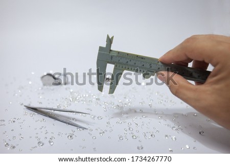 Hand holding a diamond with vernier caliper, tweezers and a pile of diamond. Worker measures the diameter of gems on a jewelry scale isolated on white background.