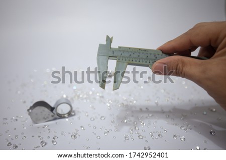 Hand holding a diamond with vernier caliper, tweezers and a pile of diamond. Worker measures the diameter of gems on a jewelry scale isolated on white background.