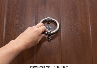 A hand holding a door knocker. Knocking on a wooden door with a metal ring shaped door knocker.