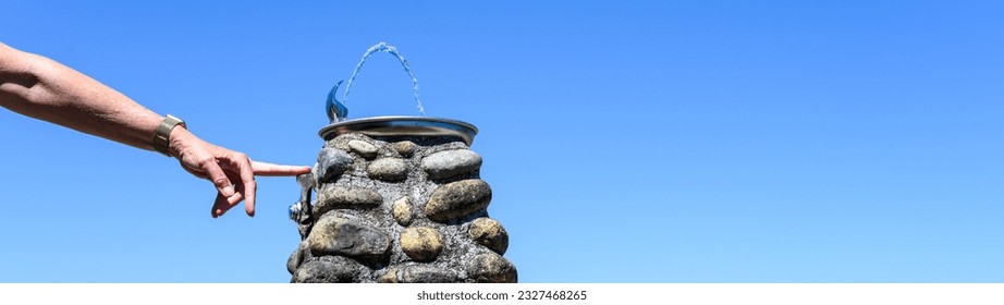 Hand holding down lever on water fountain, refreshing on a sunny summer day outside in fresh air, classic river rock and mortar pillar as base for fountain
