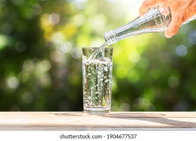 Hand holding drinking water bottle pouring into glass on wooden table on blurred green nature background
