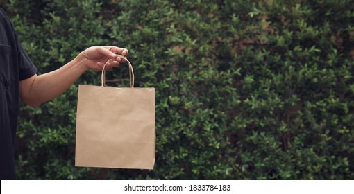 Man’s Hand Holding Empty Craft Brown Recycled Paper Bag With Outdoor Green Leaves Nature Background And Copy Space.