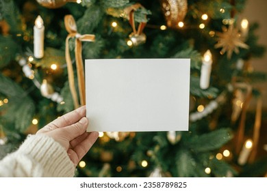 Hand holding empty greeting card on background of stylish decorated christmas tree with golden lights. Christmas card mock up. Space for text. Season greetings template