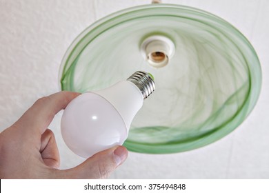 Hand Holding An Energy-efficient LED Lamp, Replacement Of The Light Bulb In The Ceiling Fixture With A Shade Of Green Frosted Glass, Close-up.