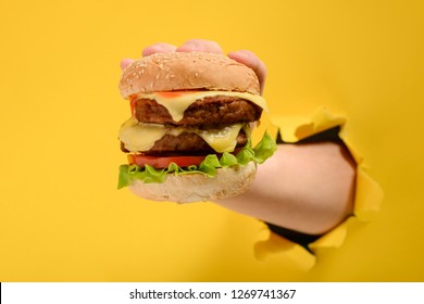 Hand holding an enormous burger through torn yellow paper background. Double beef and Cheddar cheese.