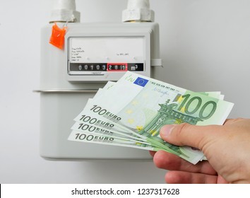 Hand Holding Euro Cash In Front Of Natural Gas Meter At Home. Symbolic Image Of A Payment For Heating In Winter, Concept. Selective Focus, Close-up.