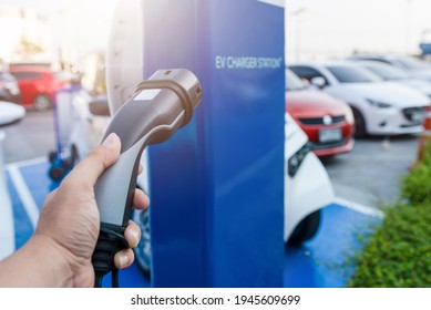 Hand Holding Ev Car Charger With EV Charging Station At The Car Park Area. Battery Charging For Automobile. Clean Energy For Sustainable Environment. Mega Trend In The Near Future.