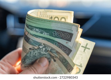 Hand holding folded stack of Polish zloty 100 banknotes with blurred car interior dashboard in background. Concept of driver earnings, cash savings, fuel budget and automotive financial transactions. 