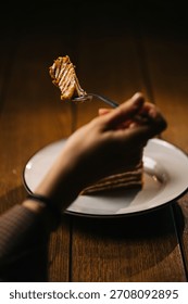 Hand holding a fork with a bite of layered cake over a plate. Warm moody dessert scene on wooden table with copy space.