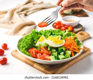 Hand Holding a Fork with Cherry Tomato. Healthy Salad of Fresh Green Coral Lettuce, Cherry Tomato, Cucumber, Carrot and Egg on a Cutter Board and White background
